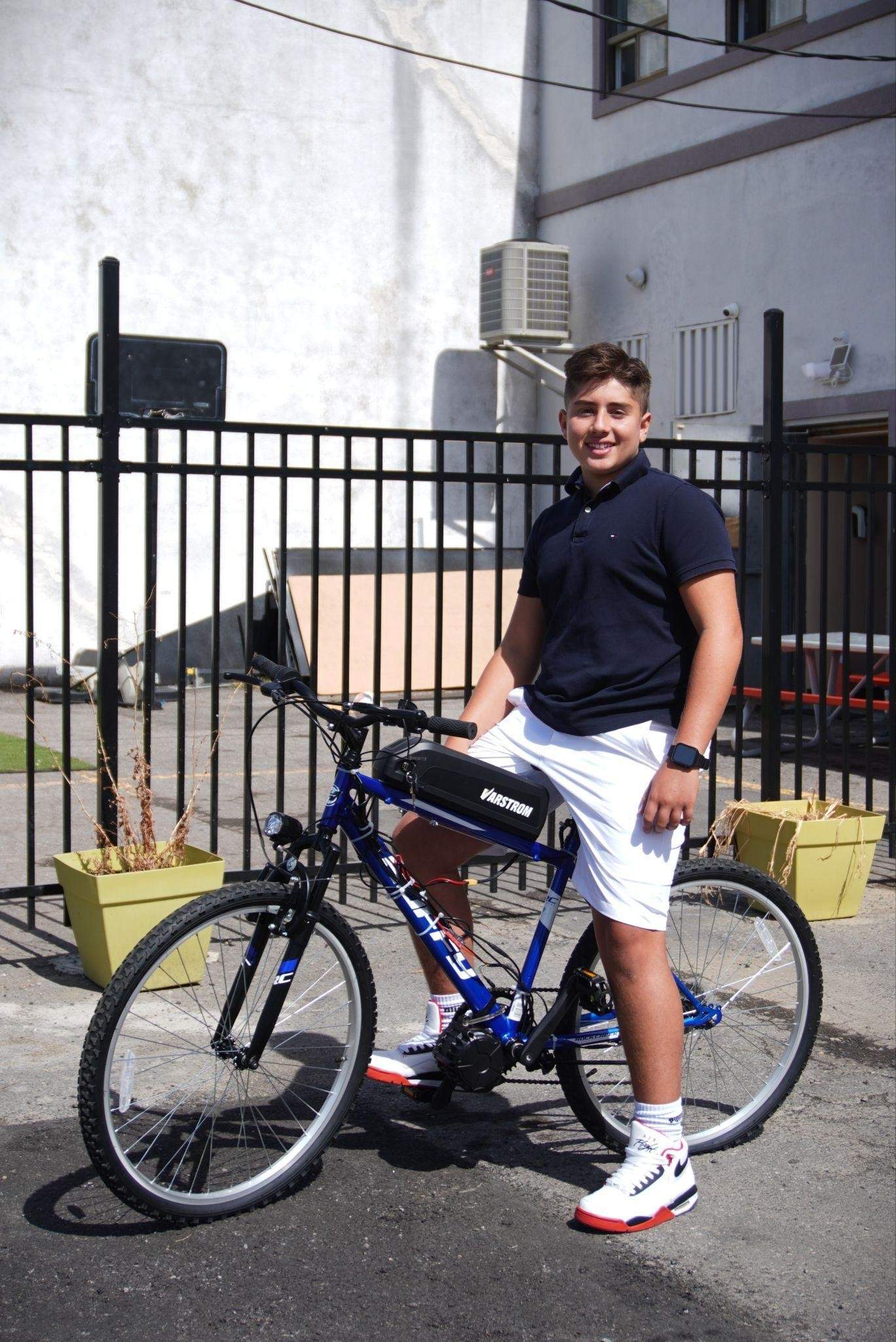 Teen engineer Marc-Anthony Mourad sitting upon one of the e-bikes built by participants of the summer e-bike workshops outside the Centre Lavallois de Ressources Éducatives et Culturelles (LREC) (Photo: Matthew Daldalian, Newsfirst Multimedia).
